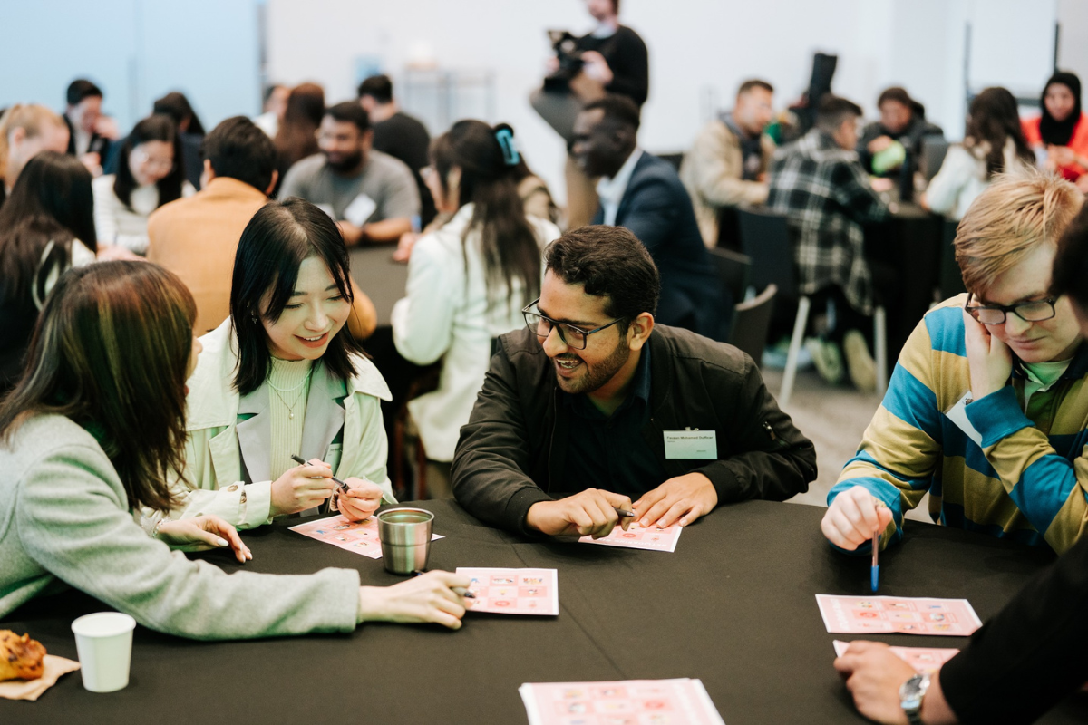Group of young adults sitting at tables, smiling and playing a game together during an indoor event with many people in the background.